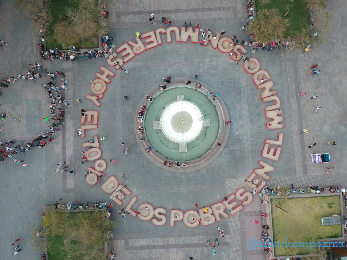 Persone riunite in una piazza attorno a una fontana centrale e a del testo.